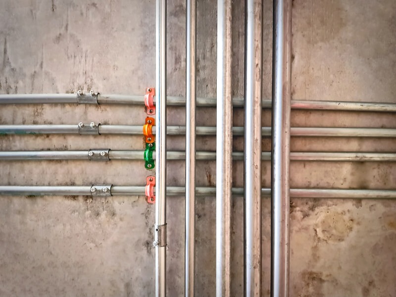 A network of parallel vertical and horizontal metal utility pipes secured to a concrete wall with colorful brackets, demonstrating professional water supply line services.